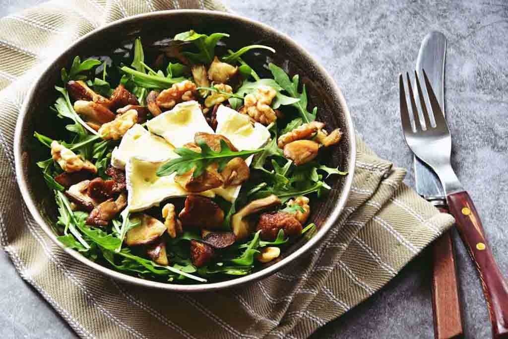 A salad plate containing brie cheese, arugula leaves, walnuts and dried figs displayed with a napkin, fork, and knife on a marble table