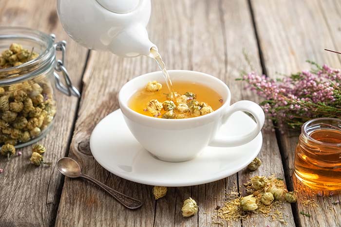 Chamomile tea poured into a white teacup alongside dried chamomile buds, fresh flowers, a jar of honey and a metal teaspoon on a wooden table
