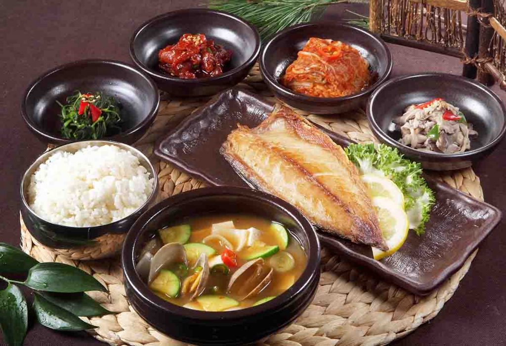 A bowl of rice, a bowl of soup, fried fish, kimchi, and other traditional Korean dishes are served on a rattan placemat
