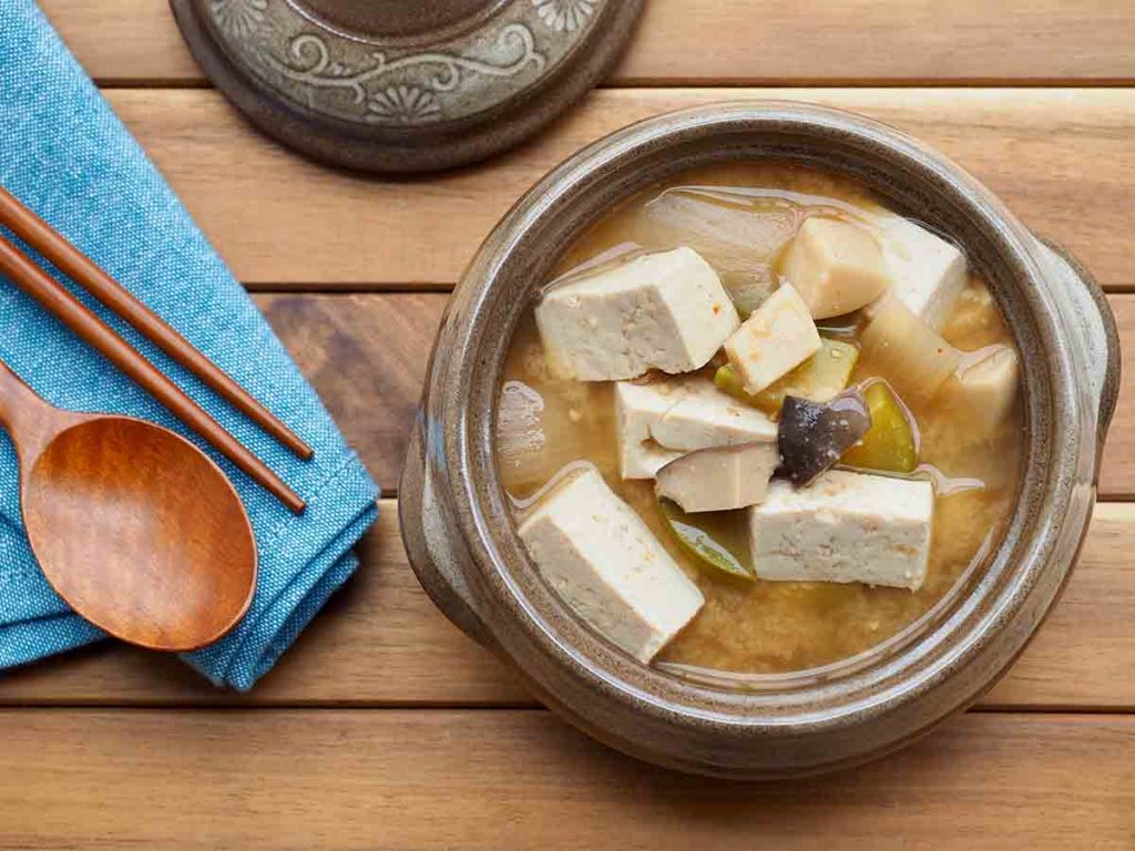 A bowl of doenjang jjigae with tofu topping alongside a wooden spoon and fork