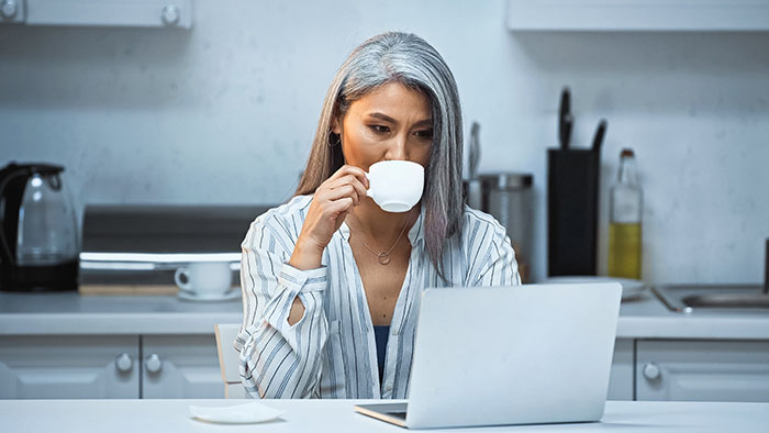 grey haired asian freelancer drinking coffee while working at laptop in kitchen
