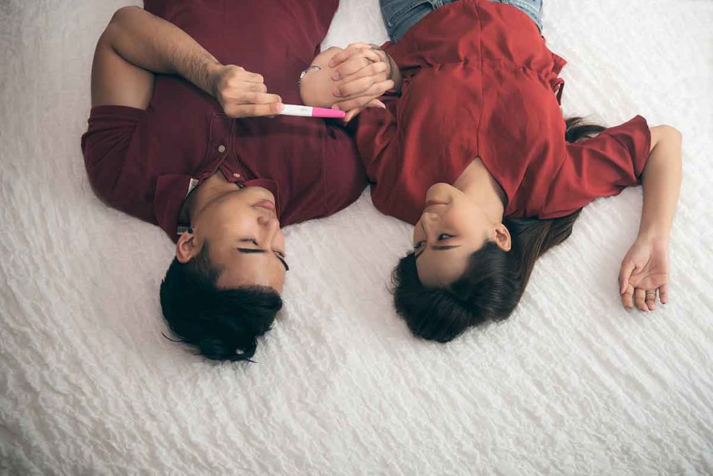 An Asian couple lie down on a bed and hold hands, while the man holds a pregnancy test kit