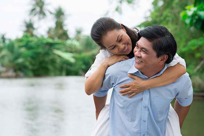 Middle-aged man giving his partner a piggyback ride as they smile at each other happily near a lake