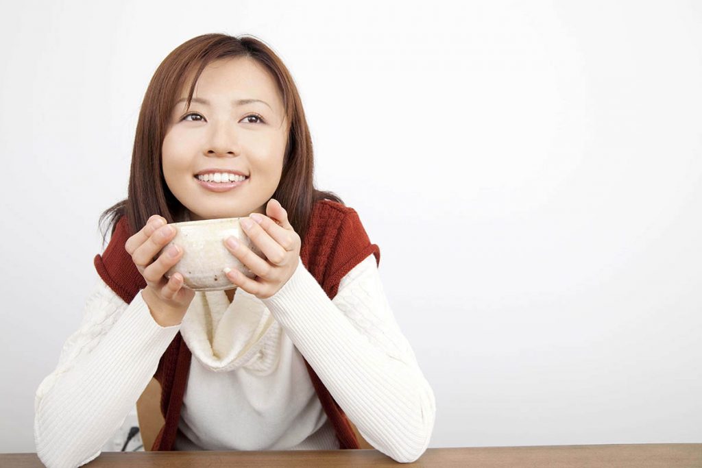 An Asian woman holds a bowl of soup, looks into the distance happily