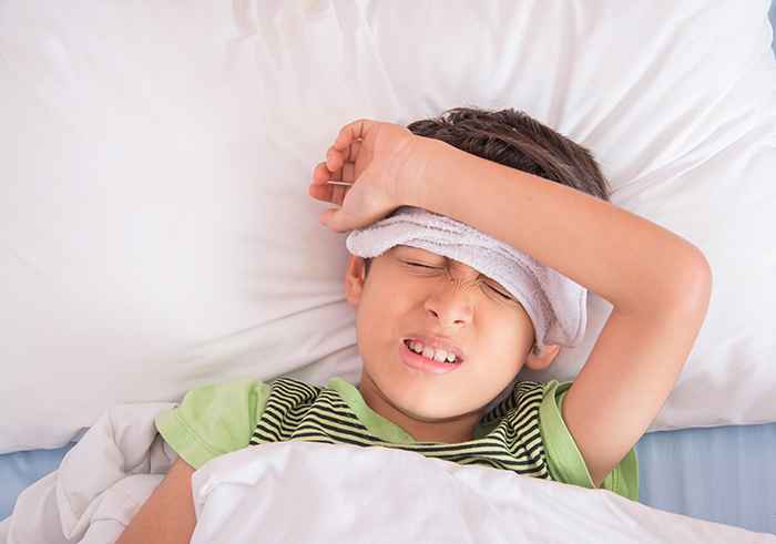 A boy in a green t-shirt lies in bed with a hand towel and his left arm on his forehead