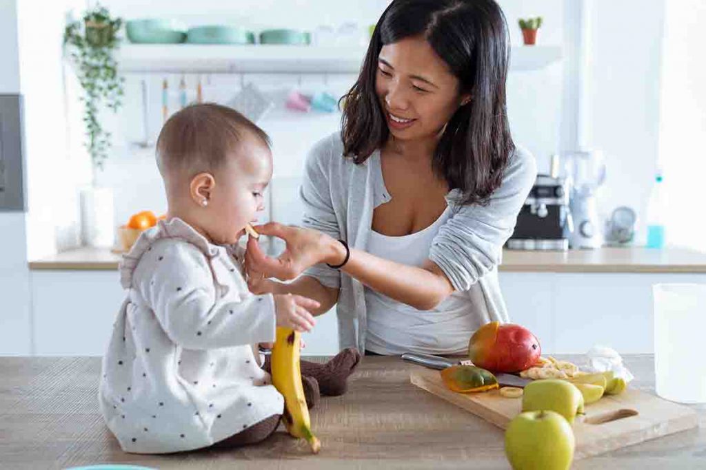 A mother feeding her baby in the kitchen with a variety of fruits on a table