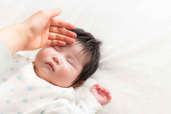 A baby sleeping on a white sheet, as their mother's hand touches their forehead to check their temperature