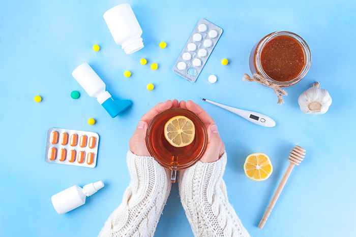 A shot taken from above of two hands around a warm cup of lemon tea, surrounded by pills, a thermometer, and a herbal drink