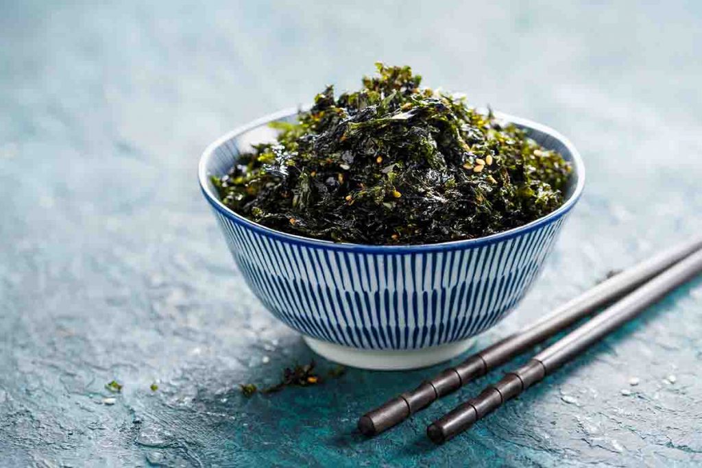 A bowl of wakame and a pair of chopsticks displayed on a flat, blue surface