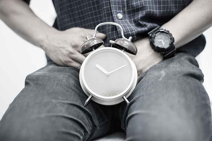 Man placing his hands near his crotch with an alarm clock supported by his inner thighs