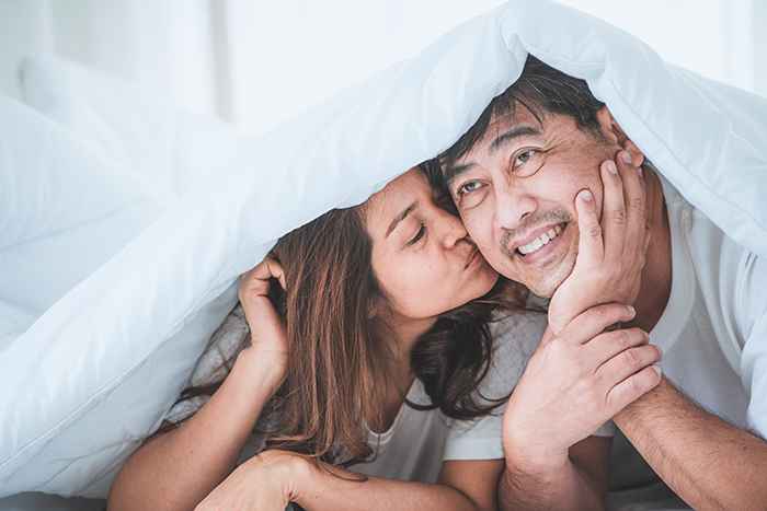 A woman kissing a man’s cheek under a bedcover