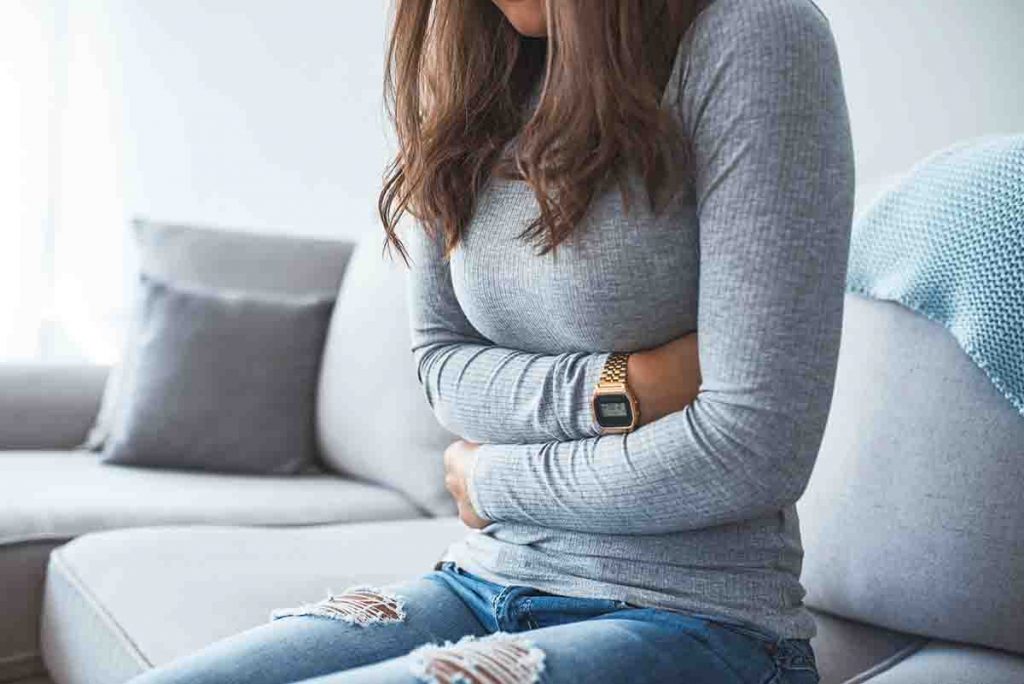 Woman wrapping her arms around her midsection in pain as she sits on a sofa