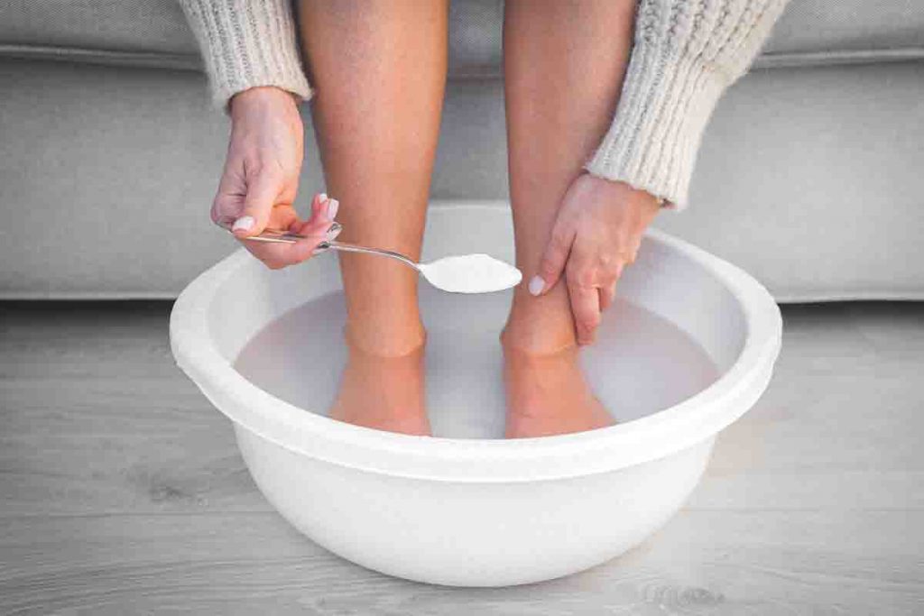 Woman soaking her feet in a tub of water as she is about to drop a spoonful of salt in
