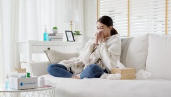 Woman with a scarf on her shoulders sitting on a sofa while blowing her nose