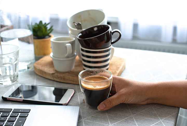 Woman holding a cup of coffee that’s next to a laptop, mobile phone and a stack of empty cups