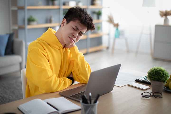 A tired man working from home, sitting in front of a laptop while holding his neck