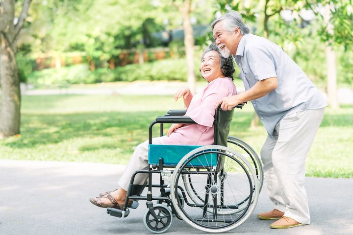 Elderly man pushing an elderly woman sitting on a wheelchair as they laugh together outdoors