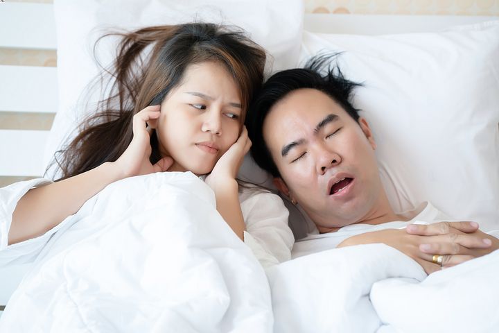 A woman closing her ears and looking at a man snoring as they both lie in bed
