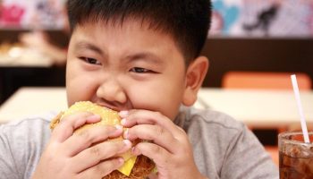 An Asian boy is eating a fried chicken burger with a glass of soda