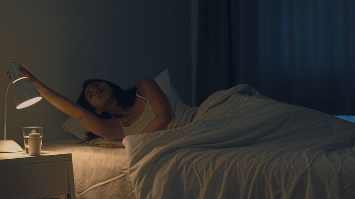 Woman stretching her right hand out to adjust the position of her bedside lamp as she lies in bed