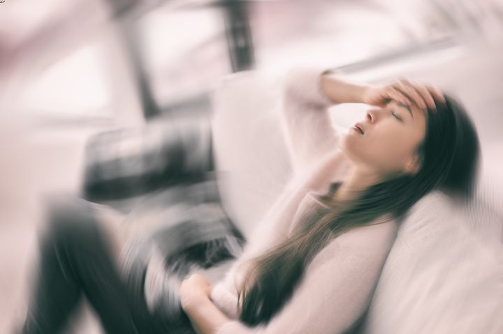 Woman with closed eyes holding a palm to her forehead as she sits on a sofa while everything around her is spinning