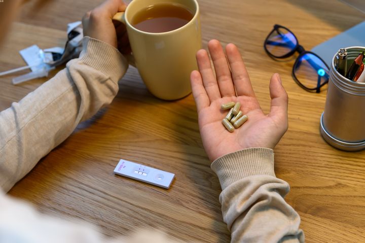Person holding cup of tea in left hand and herbal pills in right hand with a positive antigen test cartridge on the table