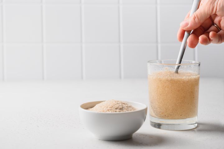 A partial view of a hand stirring a glass of fibre supplement with a spoon with a bowl of psyllium powder beside it.