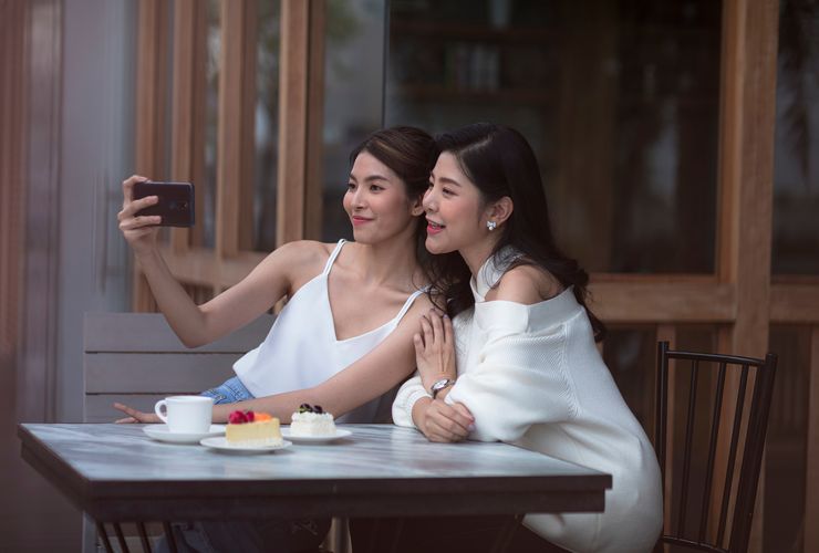 A mother and daughter posing for a selfie while sitting at a table with tea and cakes on it.