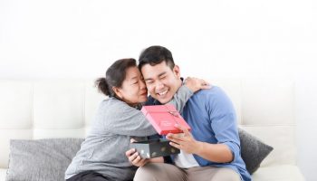 A mother hugging her happy son as he opens a gift box for her.