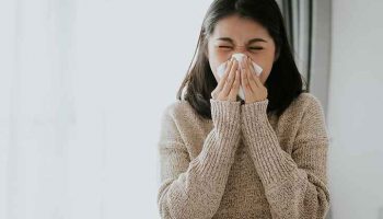 Woman in a wool sweater sneezing into a tissue that she’s holding with both hands