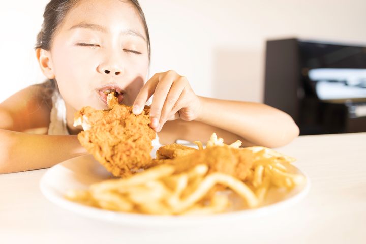 Girl biting into a piece of fried chicken with her eyes closed as a plate of various fried foods is laid out in front of her
