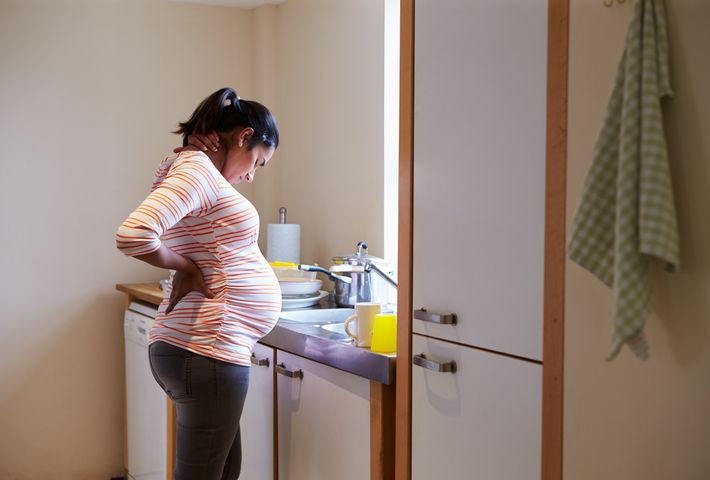 A pregnant woman standing in front of a sink while holding her back with both hands.