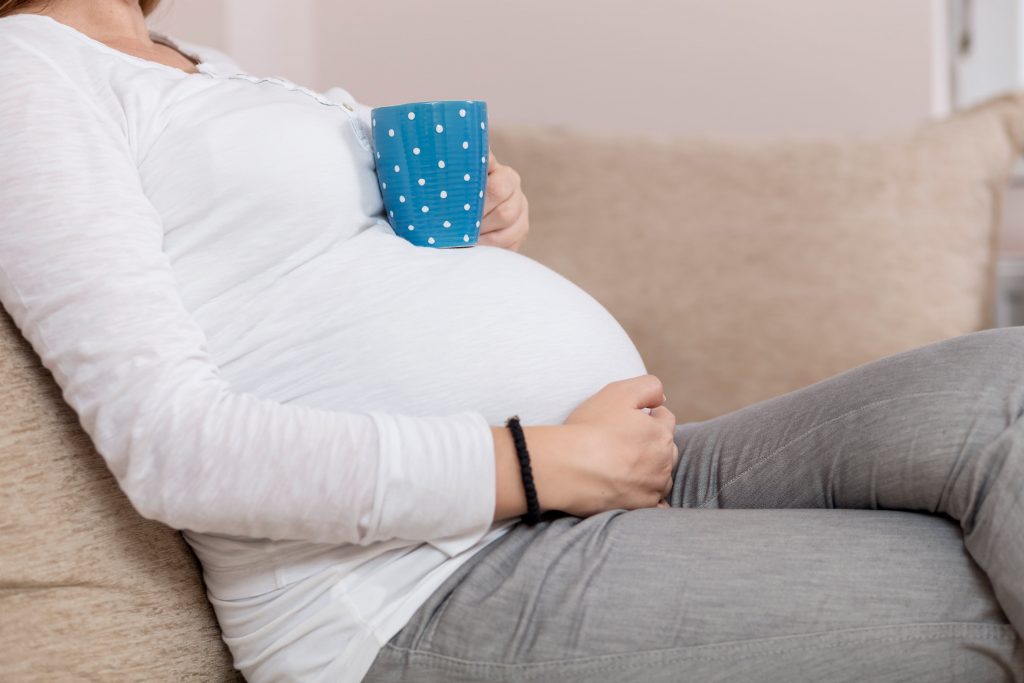 Pregnant woman sitting on a couch holding her belly and a cup of tea