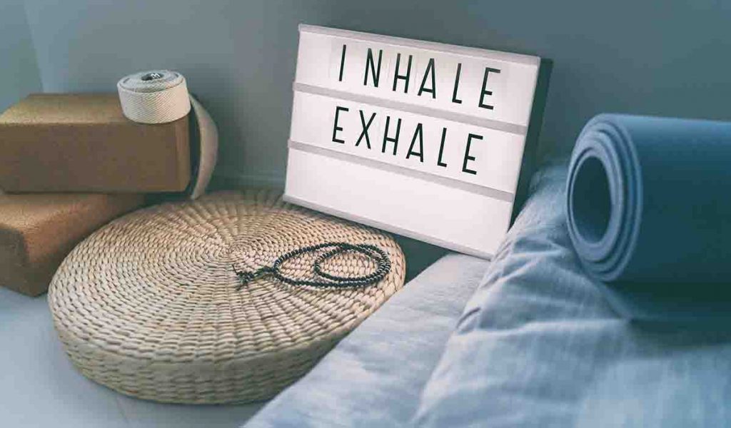 A rattan cushion, two yoga blocks, blue mattresses, and a sign that says “Inhale/Exhale”