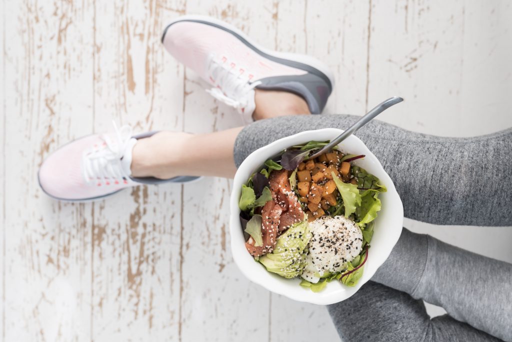 Woman in exercise clothes and walking shoes holding a bowl of avocado, protein, sweet potato, and seasonings over leafy greens