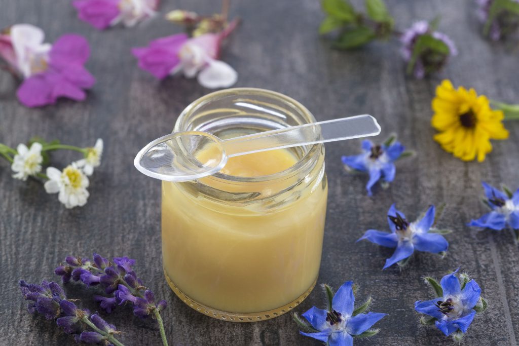 Raw organic royal jelly in a small bottle with a spoon on a background with blue, white, and yellow flowers