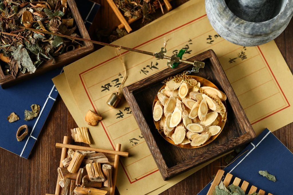 A bowl of astragalus root sits on a desk surrounded by traditional Chinese herbs