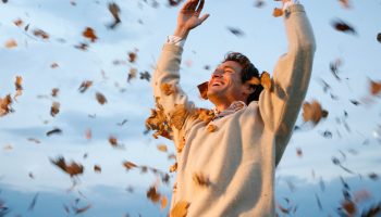 A happy man smiling widely as he raises his hands up in the air with autumn leaves around him