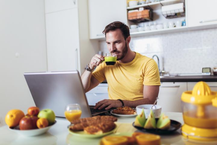 A young active man having breakfast with a cup of coffee and fruits while working on a laptop