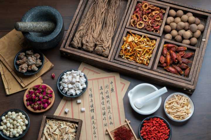An assortment of traditional chinese herbs on a dark wood tabletop wtih ginseng roots, goji berries, astragalus roots, and hawthorn berries